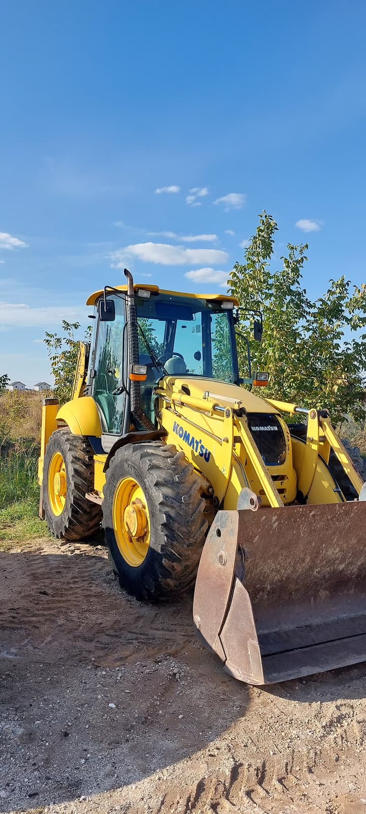 Buldoexcavator Komatsu galben cu cupă frontală, pregătit de lucru pe un teren de construcție