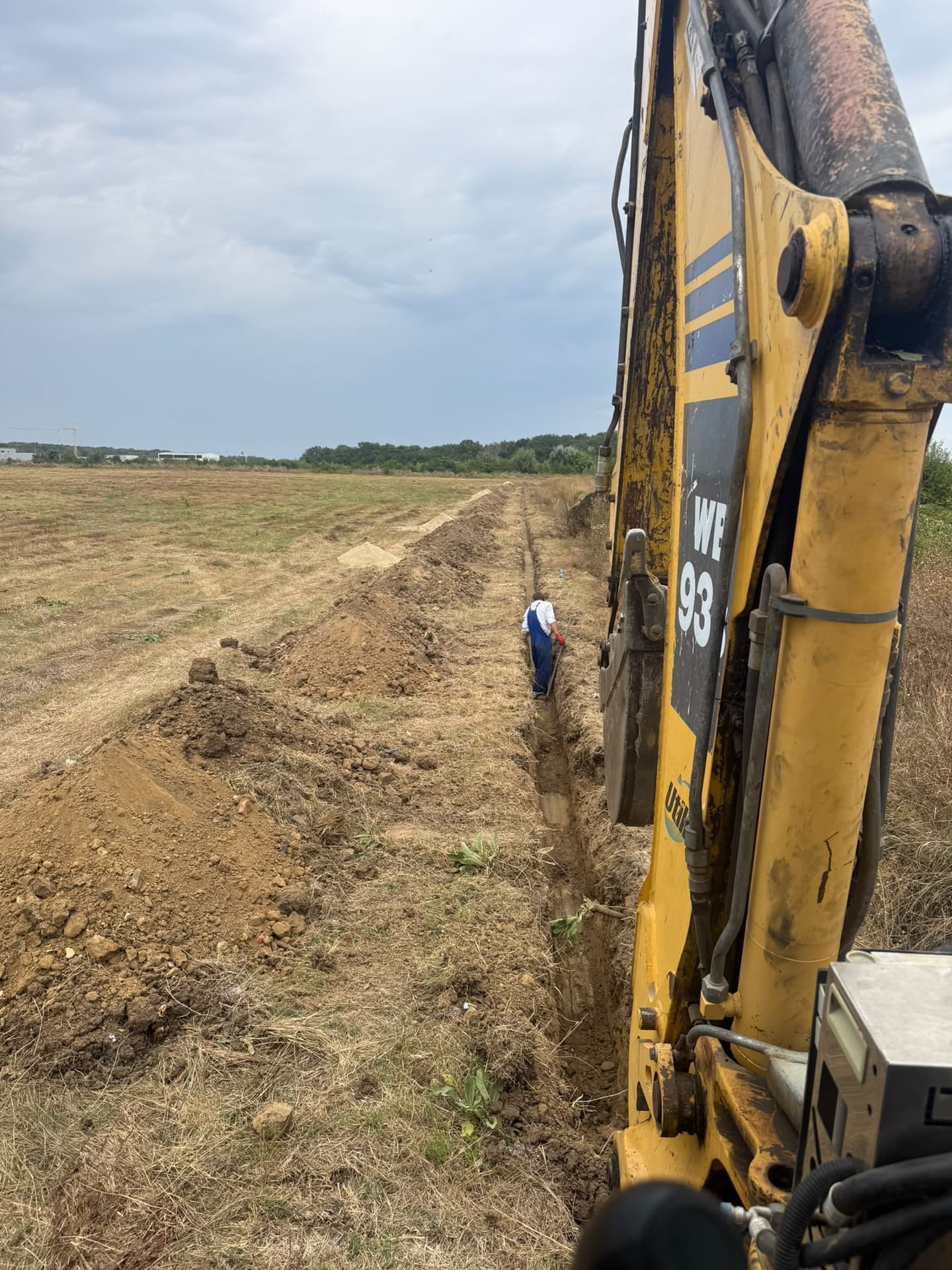 Buldoexcavator Komatsu WB93R la săparea unui șanț lung pe teren viran, cu un muncitor în șanț