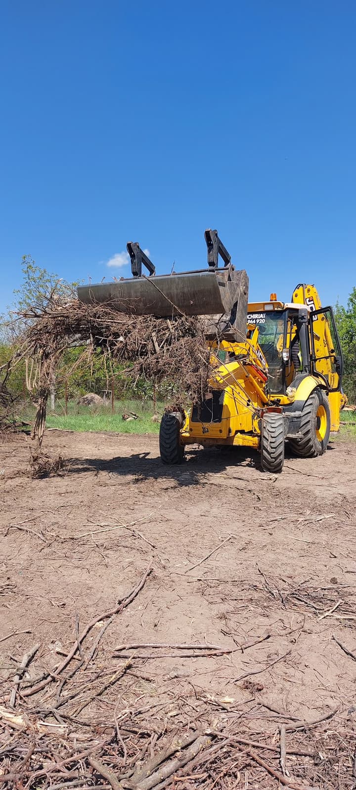 Buldoexcavator JCB cu greifer pe cupă ridică crengi și arbuști la curățarea unui teren
