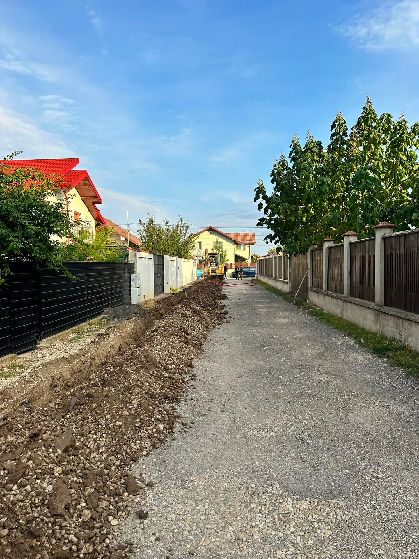 Șanț lung săpat pe marginea unei străzi pietruite, cu buldoexcavatorul la lucru în fundal